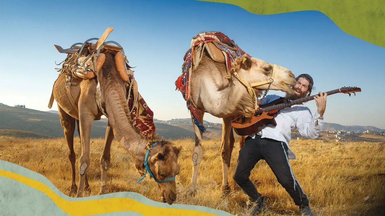 A bearded Yosef Gutman plays an acoustic bass guitar in a dry, open field as a saddled camel leans in and appears to lick or nuzzle his face, while a second camel grazes nearby against rolling hills and a clear sky.